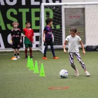 Summer Camps For Kids | Four children practicing on an indoor field. A girl dribbles a soccer ball around green cones while three children wait in the background.